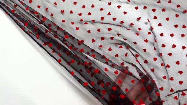 Thin translucent black mesh material displays small red velvet hearts in a repeating pattern that flows with a delicate drape for romantic fashion and decorative craft projects.