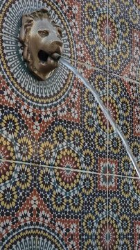 A traditional Moroccan wall fountain featuring a decorative lion head spout with a stream of flowing water, set against a vibrant mosaic tile background. 
