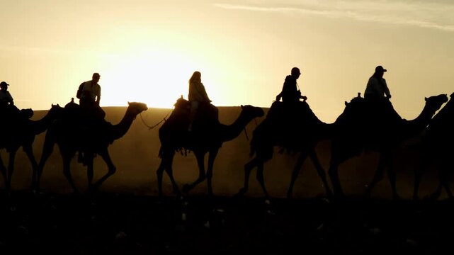 Silhouette of camel caravan with riders walking in desert against background of mountain at sunset