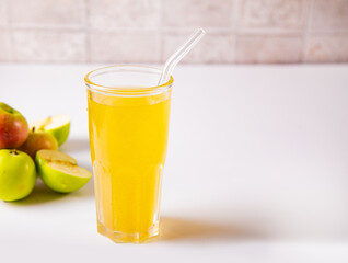 Fototapeta premium A refreshing glass of golden apple juice or cider with a reusable straw, surrounded by fresh apples and a red checkered cloth on a light background.