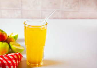 Fototapeta premium A refreshing glass of golden apple juice or cider with a reusable straw, surrounded by fresh apples and a red checkered cloth on a light background.