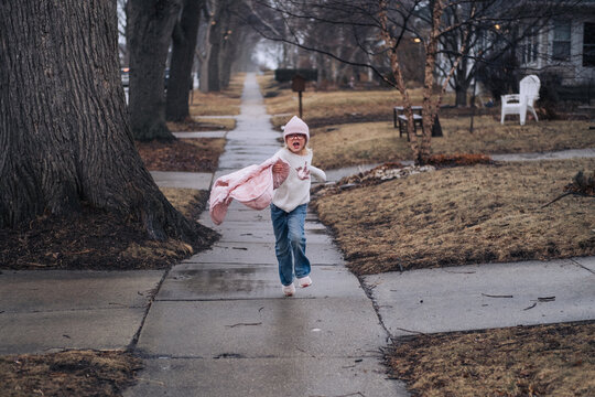 Young girl wearing a pink jacket and hat running toward the camera on a sidewalk in a residential neighborhood. Candid lifestyle moment with natural light, capturing movement, energy, and playful chil