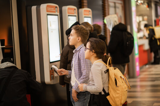 Two kids use a self-service fast food ordering machine indoor, holding a ticket and card, surrounded by people.