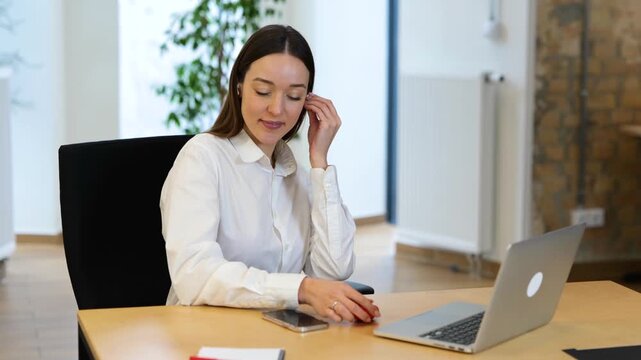 Relaxing work in a modern office. Realtime video. Caucasian beautiful female is sitting in front of her laptop and putting her earphones, and listening to music.