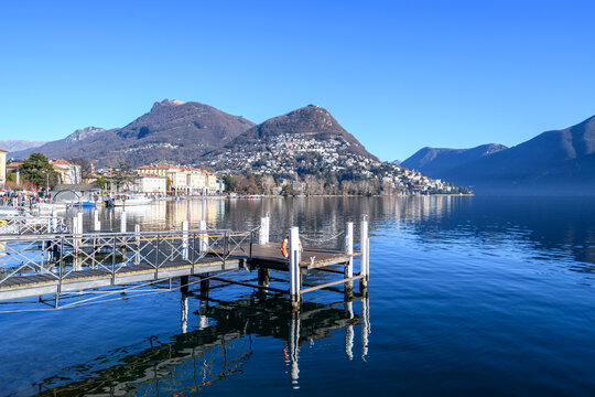 Peaceful waterfront scene in Lugano with a wooden pier, calm lake reflections, and mountain views under a clear blue sky, capturing serenity and Swiss travel charm.
