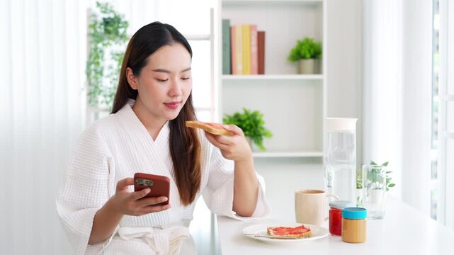 Smiling young asian woman enjoying breakfast, coffee and bread  while  browsing mobile phone for news and social media content . morning routine.