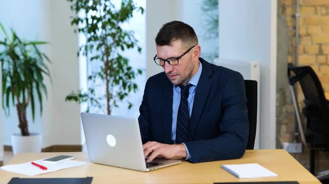 Realtime video. Caucasian male in black suit and blue shirt is sitting on his chair, typing on his computer and looking carefully at the screen of his laptop.