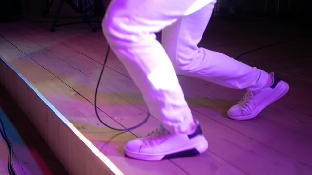 Dynamic low angle shot showcases performer wearing white trousers and matching sneakers executing rhythmic dance moves on wooden stage floor during live event under neon purple lighting.