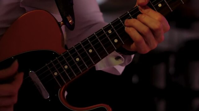 male hands strumming orange electric guitar strings featuring maple neck chrome hardware white shirt sleeve blurred nightclub background atmospheric stage lighting.