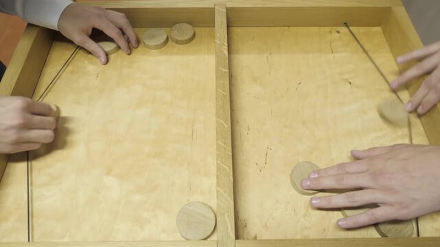 Two pairs of hands rapidly launch round wooden pucks using elastic bands across a vintage tabletop board game featuring a central divider and smooth plywood surface.