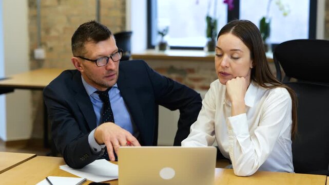 Realtime video. Caucasian man and woman re working in office. They are discussing, looking at screen of a laptop, and making notes in a notebook.