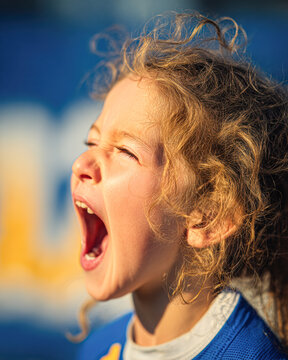 A young child with curly hair is passionately shouting or cheering outdoors in bright sunlight.