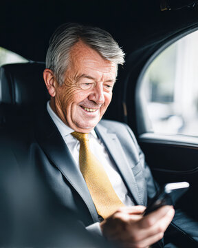 Smiling senior businessman in a suit uses a smartphone while sitting in the backseat of a car, appearing engaged and content.