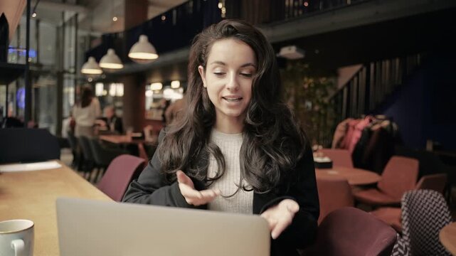 Cheerful female blogger online networking via laptop. Happy lady talking to laptop camera, explaining something in coffee shop. Workplace in cafe. Real time