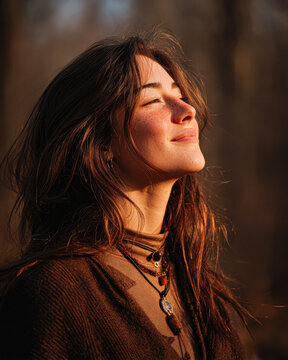 A young woman with long hair enjoys the sunlight outdoors, eyes closed, looking peaceful and content.