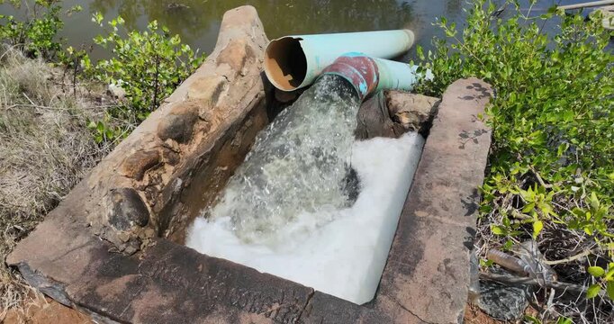 Slow motion of a rural pumping station using a diesel engine to discharge water from blue pipes, creating white foam and bubbles in a stone-lined basin.