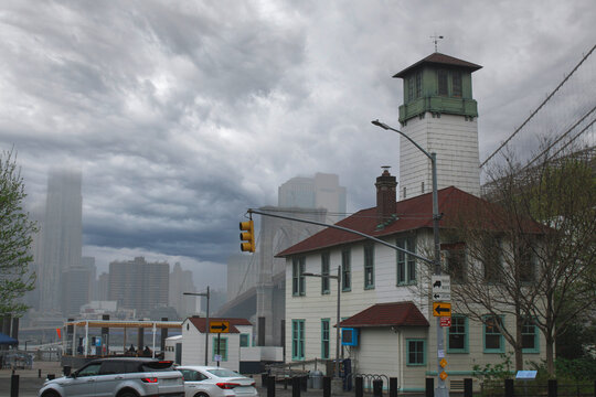 A historic white fireboat station house stands in Brooklyn Bridge Park under a dramatic, moody storm sky. The silhouette of the Brooklyn Bridge and the misty Manhattan skyline are visible behind.