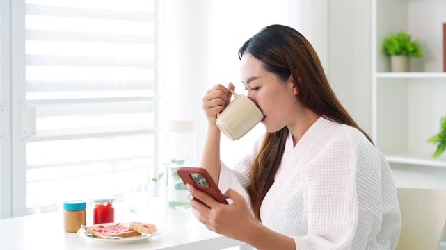 Smiling young asian woman enjoying breakfast, coffee and bread  while  browsing mobile phone for news and social media content . morning routine.