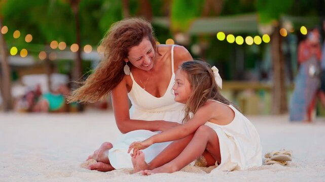 Mother with her cute little daughter in white dresses sitting on the ocean shore at sunset. Happy family on holidays. Summer vacation, travel holiday. Mother-daughter interaction