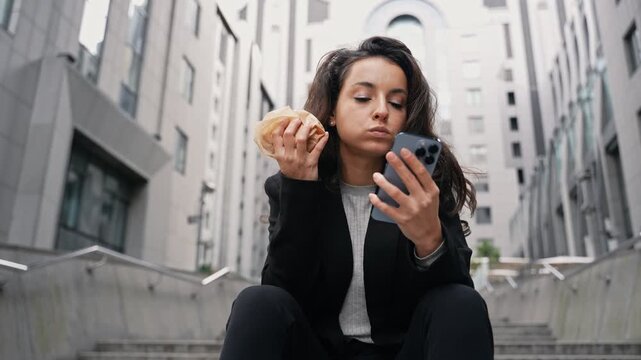 Pretty young lady working online outdoors while sitting on steps near office buildings. Woman biting a sandwich. Multitasking, lifestyle concept. Real time