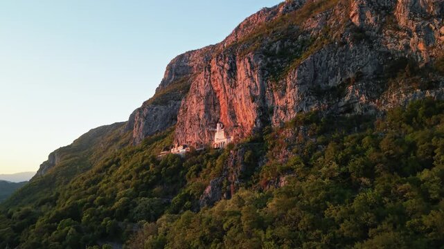 Drone shot Ostrog Monastery in the Cliff in Montenegro on sunset.  Golden hour lighting, mountains in the background.
