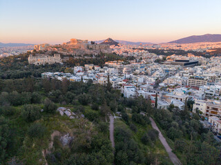 Obraz premium An Aerial Image of the Acropolis and Parthenon rising above the lush Philopappos Hill and the white buildings of Athens during the golden hour.