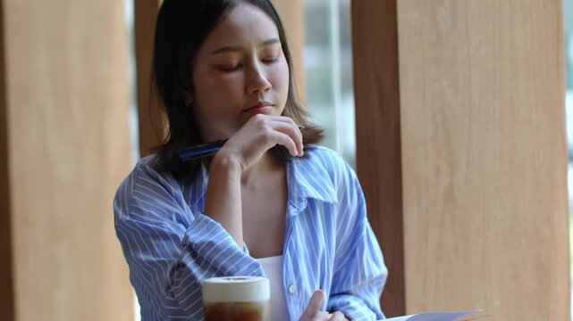 Young Asian woman writing in a notebook with a pen at a cafe, having an iced coffee with milk foam on the table, focusing on her work or studies in a peaceful atmosphere, lifestyle and education conce