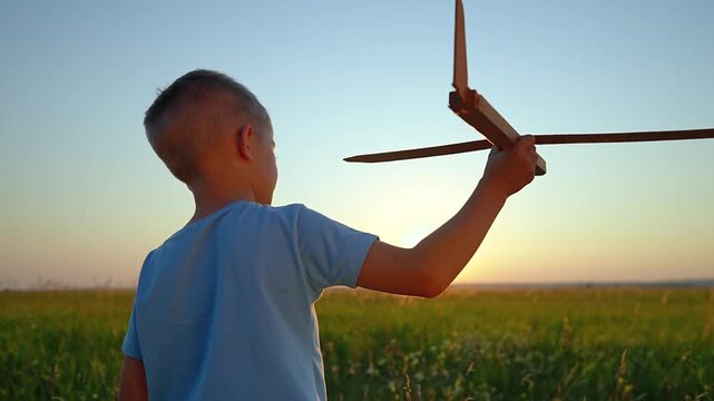Boy launches wooden toy airplane in field at sunset. Child plays with glider outdoors. Kid throws toy aircraft overhead. Boy playing with wooden plane. Child launches glider at golden hour.
