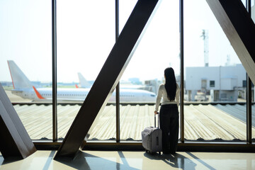 Fototapeta premium Gen Z young woman traveler waiting with suitcase watching airplane at airport window