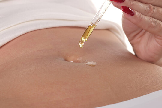 Close-up of a woman applying cosmetic oil with a dropper directly into her navel for skincare, studio shot on a white background.