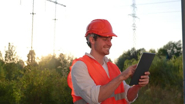 Happy male electrical engineer working outdoors while typing on the digital tablet. High voltage power lines. Energy industry concept. Real time