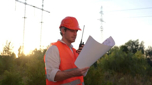 Electrical engineer talking on the walkie-talkie and holding drawing while controlling of repair of electrical high voltage pylons outdoors. Energy industry concept. Real time