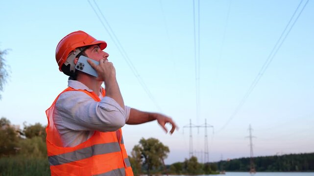 Electrical engineer in helmet checking power line while talking on the phone outdoors. Highest voltage transmission. Energy industry concept. Real time