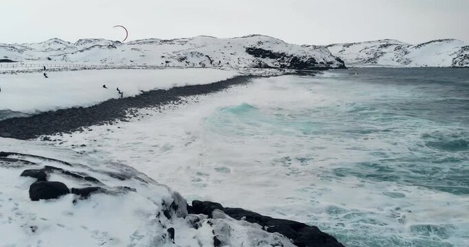 Aerial view of a snowy Arctic coastline with waves crashing and a distant kitesurfer gliding over the frozen sea.