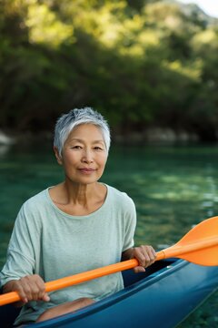Older woman holding paddle while seated in kayak on lake near forest shore