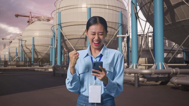 Asian Female With Her Smarphone with Spherical Propane Tanks with Fuel Gas Pipelines, She Raises Her Fist Up With Screaming Goal
