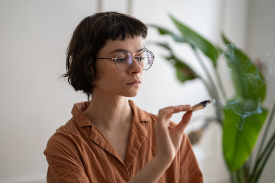 Calm woman lighting up aromatic palo santo smudging stick at home for sensory therapy and meditation, relaxed female concentrating on floating smoke in air, filling house with cozy peaceful atmosphere