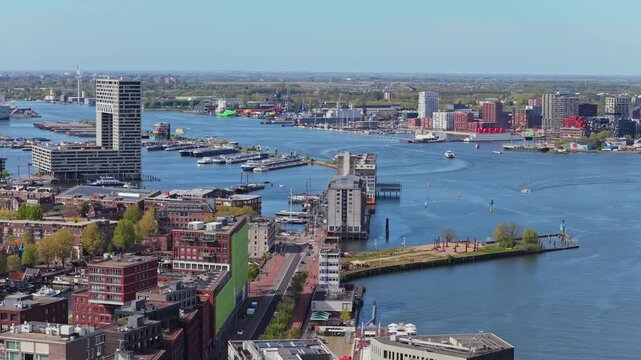 Amsterdam showing IJ river and waterfront buildings including Pontsteiger, with boat traffic moving along waterway and urban city skyline on sunny day, forward moving aerial