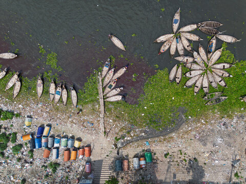 Aerial view of traditional wooden boats moored in circular patterns on the Buriganga River with colorful barrels and green vegetation in Dhaka, Dhaka Division, Bangladesh.