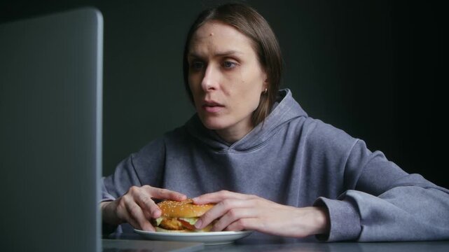 Focused woman in grey hoodie eating hamburger while working or watching content on laptop, showcasing concepts of stress eating, multitasking, and unhealthy work life balance. Unhealthy lifestyle