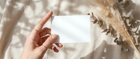 Obraz premium Female hand holding blank white business card mockup with natural shadow overlay from dried plants and pampas grass for branding presentation.