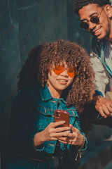 Fototapeta premium Young stylish couple sharing a fun moment looking at a smartphone outdoors, smiling in natural sunlight against a teal wall