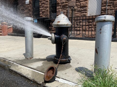 Black and silver fire hydrant with spray cap to conserve water spraying onto street on hot summer day in Harlem, New York City