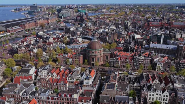 Flying over Amsterdam showing Saint Nicholas Basilica and historic canal houses with Centraal Station and urban waterways stretching towards distant horizons