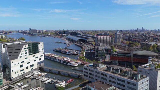 Flying over modern architecture in Amsterdam, revealing river cruise ship, Amsterdam Centraal Station, and IJ river on sunny day, establishing expansive cityscape
