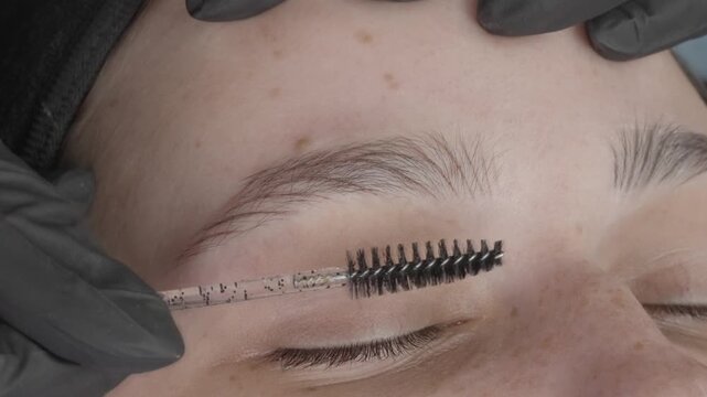 Close-up of eyebrow grooming after microblading procedure. Beauty specialist brushing shaped brows to demonstrate result. Permanent makeup concept, skincare and cosmetic treatment.