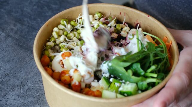 Person uses a wooden fork to mix a nutritious vegan salad with chickpeas, mung bean sprouts, and fresh vegetables in a recyclable kraft paper bowl for a healthy takeaway lunch. Vegan food concept.