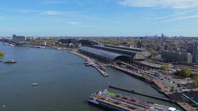 Scenic survey of Amsterdam Centraal station and IJ river, featuring city skyline, bustling traffic, boat transit on a bright, sunny day in Holland's capital