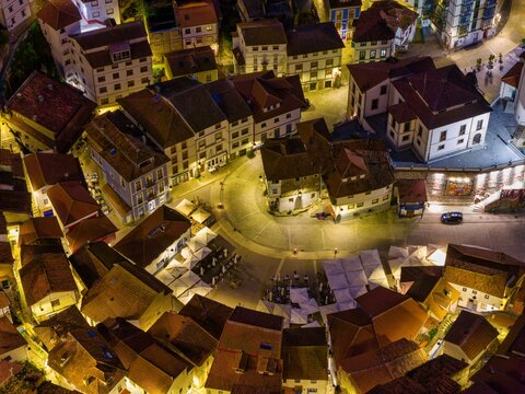 Aerial view of the illuminated Plaza de la Marina at night with traditional colorful houses and outdoor cafe umbrellas in Cudillero, Asturias, Spain.