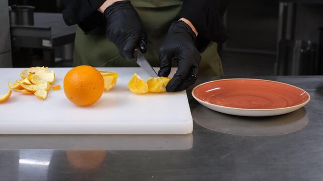 Male chef in professional kitchen slicing orange into wedges and arranging on plate

